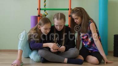 三个女生在体育课上休息时使用手机。 十几岁的女朋友坐在地上看着智能手机