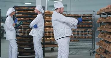 面包店里的一群面包师站在满架的新鲜烘焙面包旁边，主面包师用一个面包机拍照
