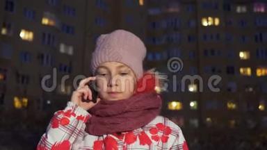 晚上打手机的少女走在冬天的大街上。 少女在冬季街头谈论智能手机