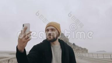 快乐年轻兴奋的白种人博客男子拍摄智能手机自拍视频史诗山圣米歇尔城堡慢动作。