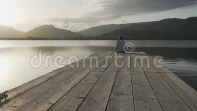 一位从背后看到长发的年轻女子在夕阳下坐在一座古老的木墩上看着湖面