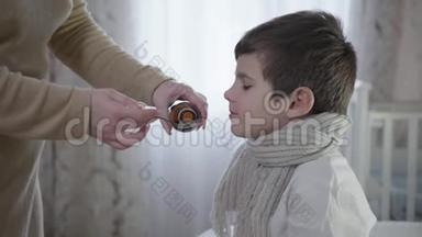 照顾孩子，家长把咳嗽糖浆倒入勺子里，让生病的孩子坐在床上吃药