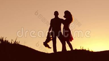 恋爱中的情侣在自然日落和接吻时跳舞剪影。 爱的男人和女人与狗跳舞剪影缓慢