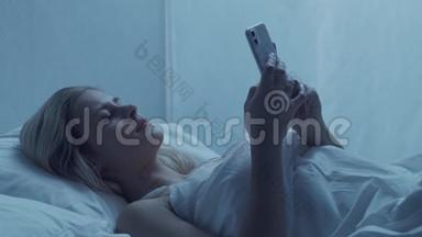 年轻的女人<strong>晚上</strong>躺在床上拿着智能手机。 美丽的金发女孩休息。 <strong>卧室</strong>里的暮色