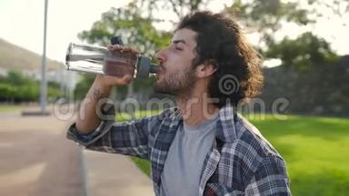 一个穿着休闲服的年轻人从公园里可重复使用的<strong>玻璃</strong>瓶里喝水的多利<strong>照片</strong>