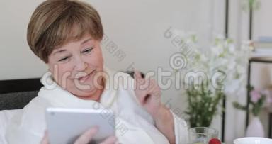 快乐成熟的女士挥手<strong>购</strong>买和吹吻，同时完成视频通话平板电脑。 在家抢劫录像的老妇人