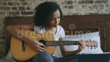 年轻的卷发混血女孩集中学习弹吉他使用笔记本电脑坐在家里的床上