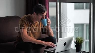 英俊的年轻人一边喝咖啡一边用笔记本电脑，坐在家里的沙发上，窗前。