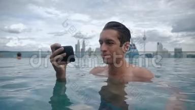 年轻人在游泳池里放松，在城市背景下摩天大楼屋顶上的智能手机上<strong>观看视频</strong>
