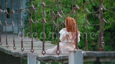 迷人的可爱的年轻女子，一头火红的头发，坐在河上的吊桥上，梳着辫子