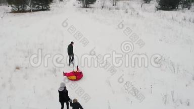 快乐的一家人在冬天的田野上玩耍-父亲骑着他的儿子在充气雪橇上，他的妻子和女儿在跑步
