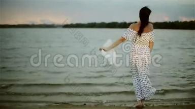一位穿着<strong>夏日</strong>长裙的中年妇女站在海滩上，脱下<strong>帽子</strong>，享受着风。