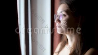 一个戴眼镜的年轻女子望着窗外，期待着来自一个黑暗的房间