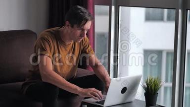 英俊的年轻人在家坐在窗前的沙发上用笔记本电脑。
