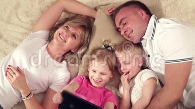 妈妈，爸爸，女儿和儿子在柔软的毯子上休息，看着装着小女儿的平板电脑。 快乐的人