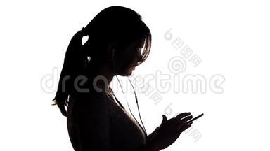 一个年轻快乐的女人戴着大耳机听音乐，拿着智能手机的剪影，女孩在白色的隔离巴上跳舞