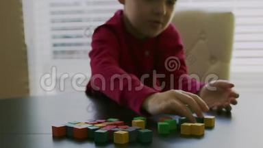 穿着红色毛衣的小男孩正在玩窗户前桌子上五颜六色的木块。 特写录像。