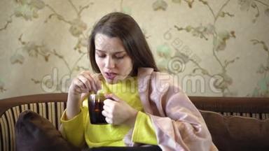 一个漂亮体贴的女孩坐在沙发上喝茶。 她肩膀上有棕色的毯子。 妇女