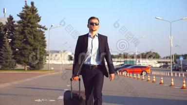 英俊的年轻商人戴着太阳镜走在城市街道上，把手提箱拉在轮子上。 自信的成功人士