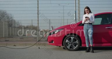 一位女士拿着电话站在她红色的电动车旁，等着车充电。连接的充电器插头