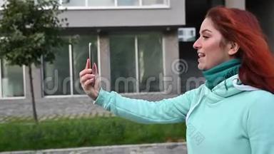 一个年轻的红发女人走在街上，在手机上录制一段自拍视频。 一个穿运动衫的漂亮女孩