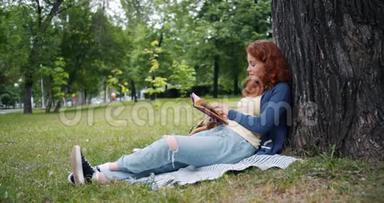 美丽的年轻女子<strong>坐在</strong>公园<strong>树下</strong>的毯子上看书，微笑着