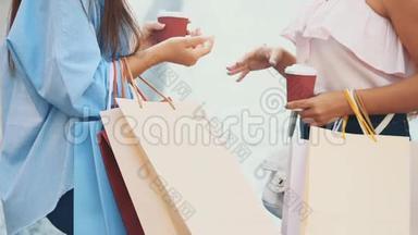 两个有魅力的年轻女人在一条带着咖啡杯的夏日街道上散步和交谈。 关上购物袋。 作物