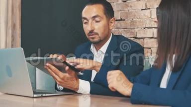 两个穿着深蓝色西装的商人正在笔记本电脑和平板电脑上工作，坐在咖啡店里。 作物。 快关门。 副本