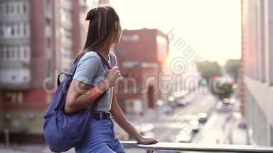 幸福微笑的年轻女子穿着休闲装，小背<strong>包</strong>坐在阳台的<strong>栏</strong>杆上