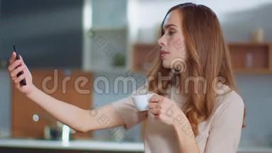 女人在家里的智能手机上自拍照片。 女孩在厨房喝咖啡