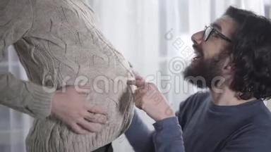 快乐的白种人男人听着婴儿<strong>撞击</strong>和亲吻他怀孕的妻子`肚子。 戴眼镜的大胡子在等