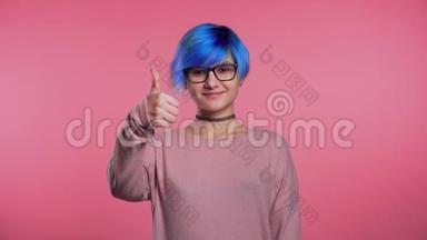 年轻的朋克不寻常的女人，蓝色的头发，竖起大拇指，在粉红色的背景上签名，微笑着对着镜头。
