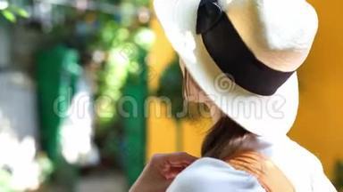 一位戴着草帽、白衬衫、微笑着对着镜头的美丽旅游妇女站在明亮的橙色墙上