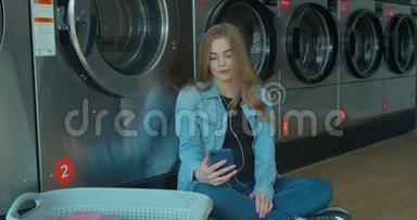 年轻<strong>的女人坐在地板上</strong>等待洗衣服，在自助洗衣店里使用智能手机。