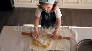 一个穿着厨师帽和围裙`可爱的小女孩在家里卷着一个擀面杖面团。