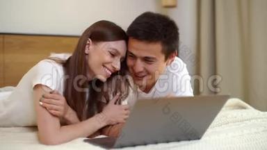 年轻快乐的白种人男人和女人躺在床上，说话，用笔记本电脑