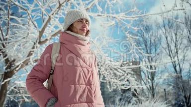 慢镜头，美丽的微笑着的年轻女子，穿着温暖的粉色派克大衣，在阳光灿烂的白雪冬日公园里散步