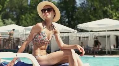 适合年轻的比基尼女士戴帽子坐在游泳池边缘的桌椅和日光浴。