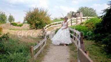 4K慢镜头视频：喜笑颜开的新娘穿着白色长裙走在乡间长木栅栏的小路上