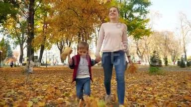 4k视频快乐微笑的妈妈和小儿子在秋天公园的草地上走着，上面覆盖着黄叶