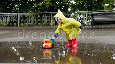 雨后，穿着橡胶靴和雨衣的小学步男孩在大水坑里与玩具卡车相撞的慢动作视频