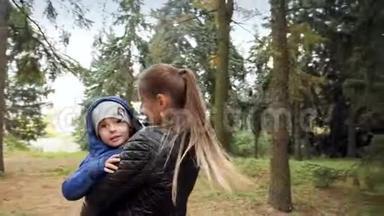 在秋日公园的高树下，一位微笑的年轻妈妈拥抱并拥抱她的小儿子