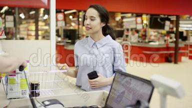 在<strong>超市</strong>里，一位美丽的女士在<strong>收银</strong>台前等着，而<strong>收银员</strong>则在她的购物篮里扫描挑选的商品，微笑着