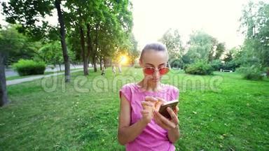 在<strong>日</strong>落城绿色公园背景下，戴着粉红色眼镜的年轻微笑女子用<strong>手机</strong>慢动作