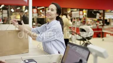 漂亮的黑发女顾客拿着智能手机，谢谢收银员，微笑着，<strong>从收银台</strong>拿着纸购物袋