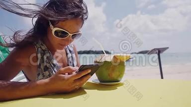 在海滩上，年轻的黑发女人戴着太阳镜，用手机，喝着<strong>椰子</strong>做的<strong>鸡尾酒</strong>