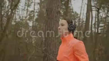 女人在公园慢跑。 自信专注的女运动员戴着耳机和橙色夹克在冬季森林的早晨跑步。 P.