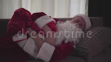 穿着圣诞老人服装，睡在沙发上，拿着带礼物的包，疲惫不堪的高年级考卡西亚人。 精疲力竭的老人
