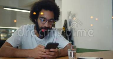 一个30多岁的大胡子，他用智能手机，坐在咖啡馆里微笑。 戴眼镜和T恤衫的快乐男人在聊天