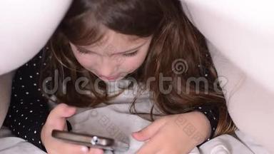 小可爱的女孩在床上的被子下玩手机